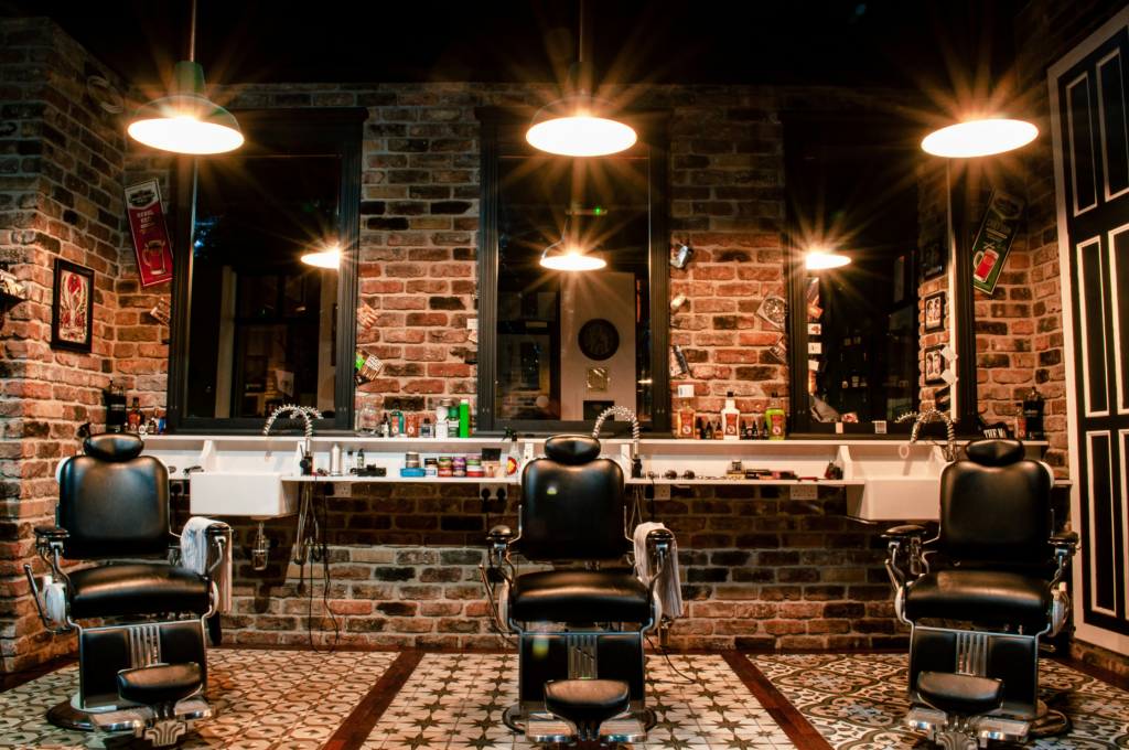 A set of three barbershop chairs facing the camera with a brick wall and three tall mirrors in the back.